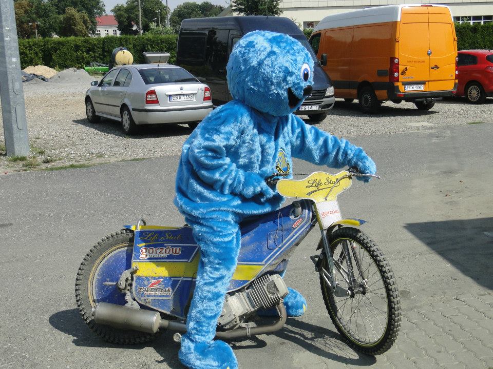 Na fotografii widnieje maskotka w postaci stworka o imieniu Staluś pozujący do zdjęcia na profesjonalnym motocyklu żużlowym